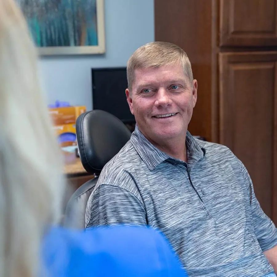 Man in gray polo shirt smiling while sitting in office chair talking to a person in blue attire.