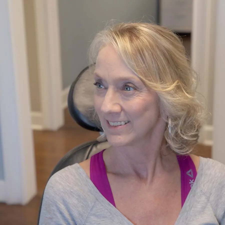 Smiling middle-aged woman with blonde hair wearing casual workout clothes sitting in a chair indoors.