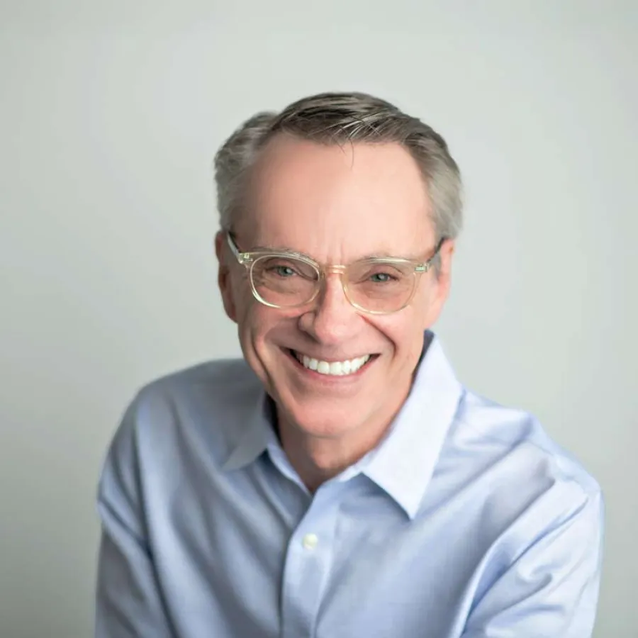 Smiling middle-aged man wearing glasses and a light blue button-up shirt against a plain gray background