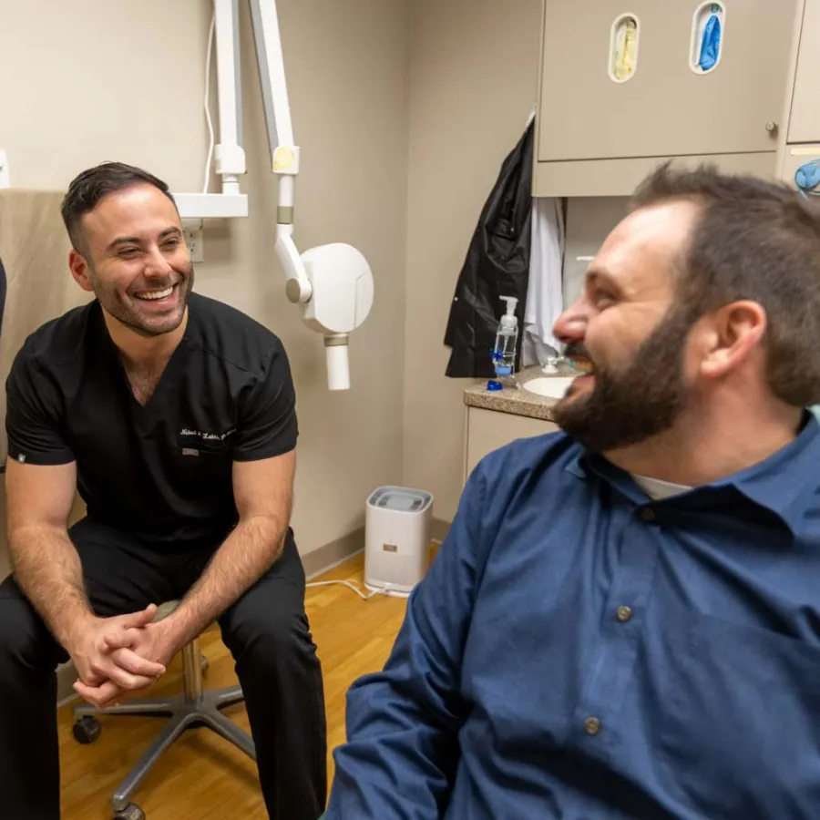Smiling dentist in black scrubs converses with male patient in a clinic room with dental equipment.