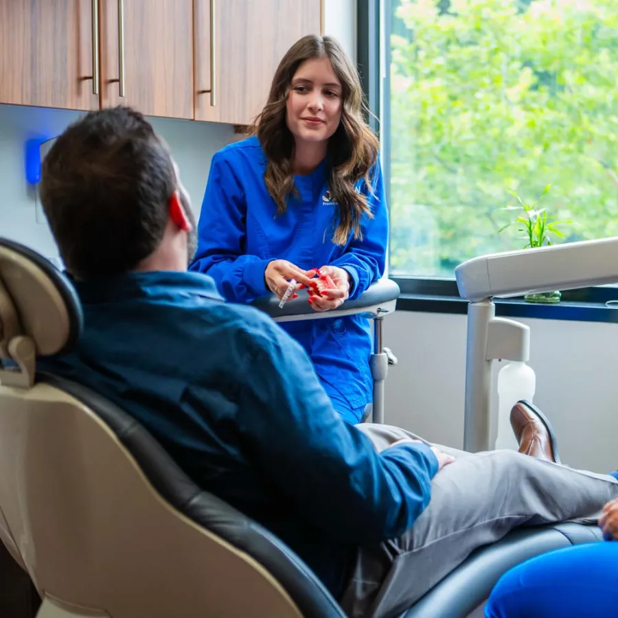 Dental hygienists in blue scrubs consulting a male patient reclining in a dental chair near a large window.
