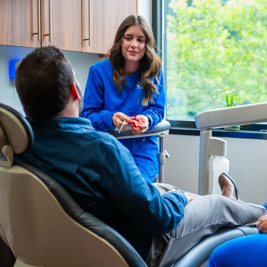Dental hygienists in blue scrubs consulting a male patient reclining in a dental chair near a large window.
