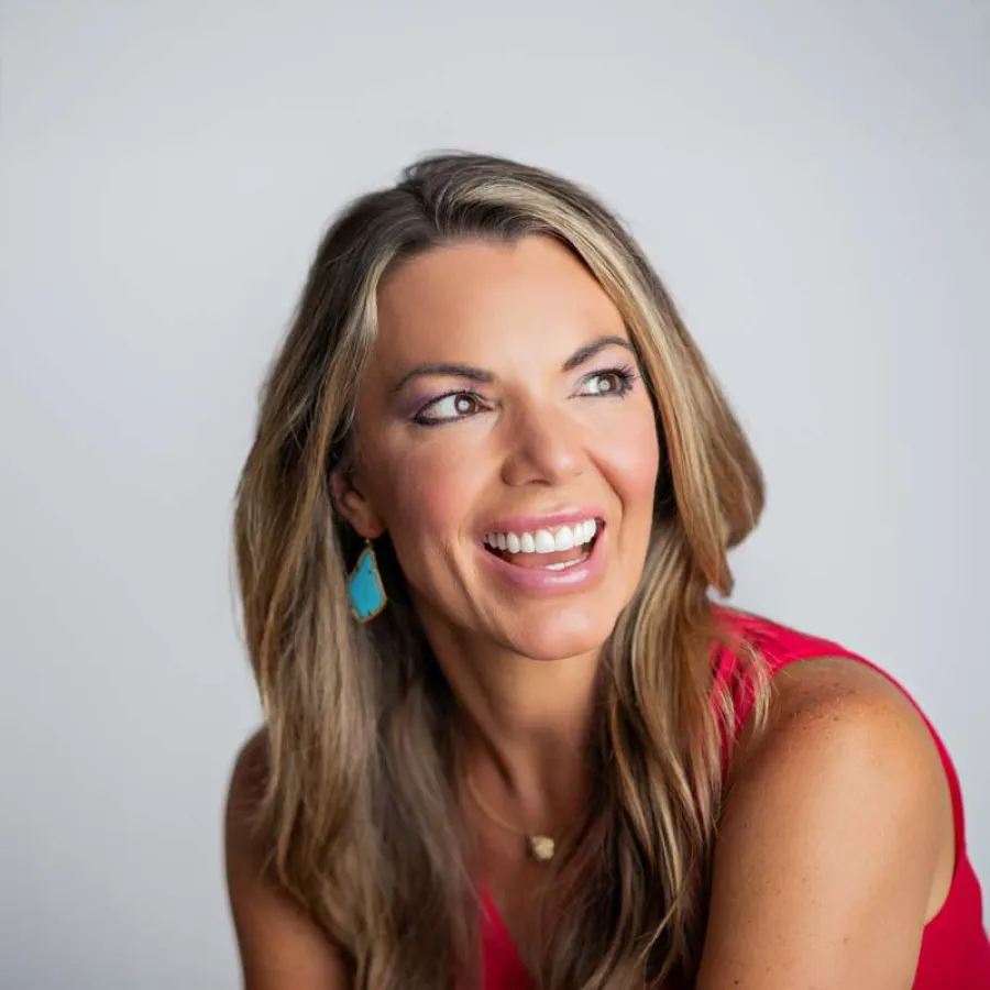 Smiling woman with long blonde hair wearing a red top and turquoise earrings looking to the side.