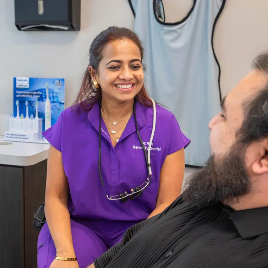 Smiling female dentist in purple scrubs talking with male patient in dental office setting.