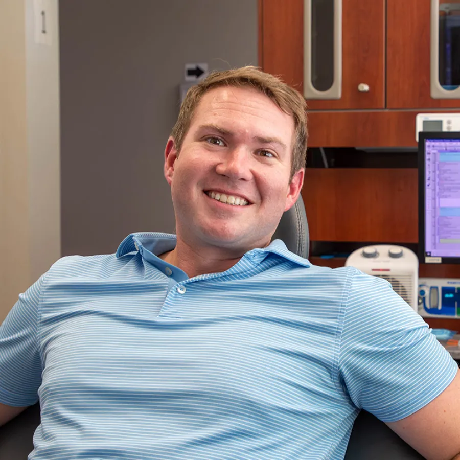 Smiling man in a light blue striped polo shirt sitting in a chair in an office setting with computer and wooden cabinets.