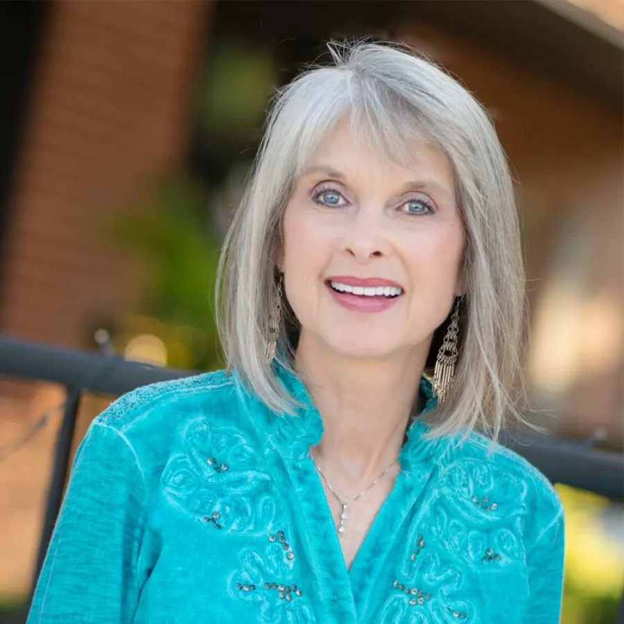 Smiling elderly woman with gray hair wearing a turquoise embroidered blouse and dangling earrings outdoors.