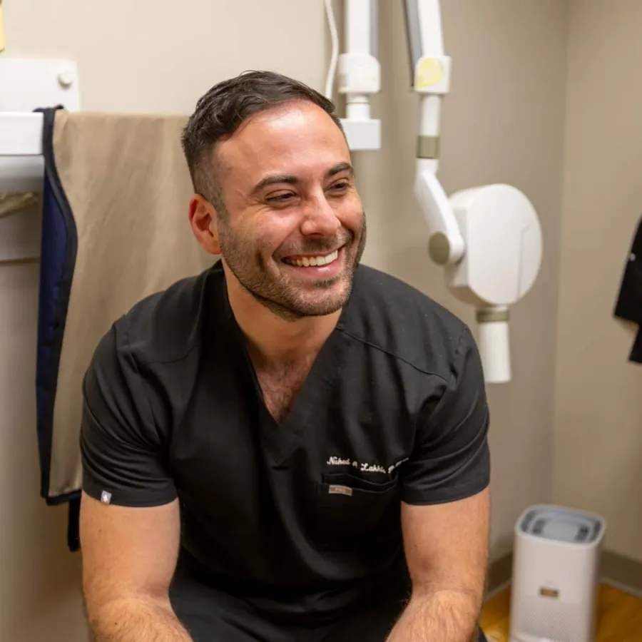 Smiling male dentist in black scrubs sitting in dental office with equipment and sink in the background