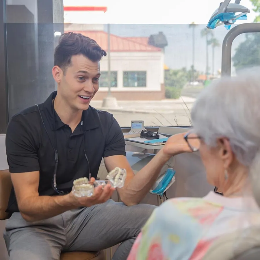 Young male dentist consulting with elderly patient in modern dental office with equipment and large window.