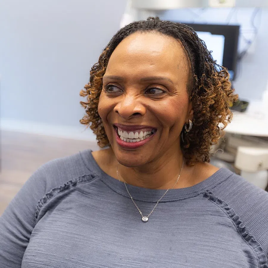 Smiling woman with curly hair and silver necklace sitting indoors wearing a gray top