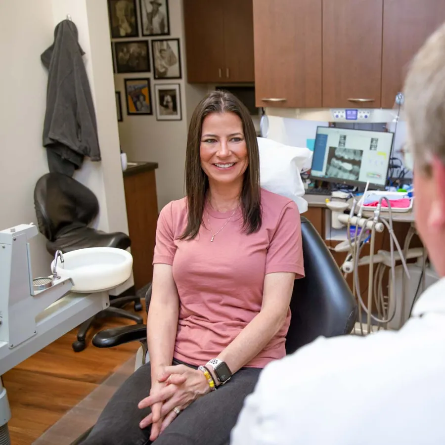 Smiling woman in pink shirt sitting in dental office talking to dentist with dental equipment in background