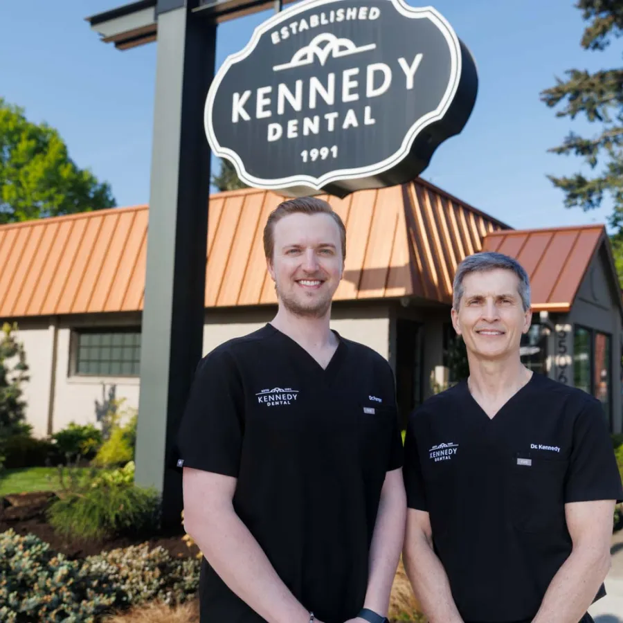 Two dentists in black Kennedy Dental scrubs standing outside their dental clinic under the clinic sign on a sunny day