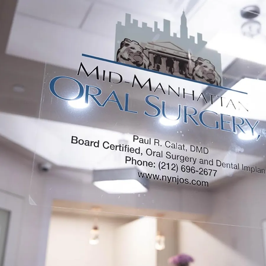 Glass door sign for Mid-Manhattan Oral Surgery featuring contact details and logo with city skyline and lions.