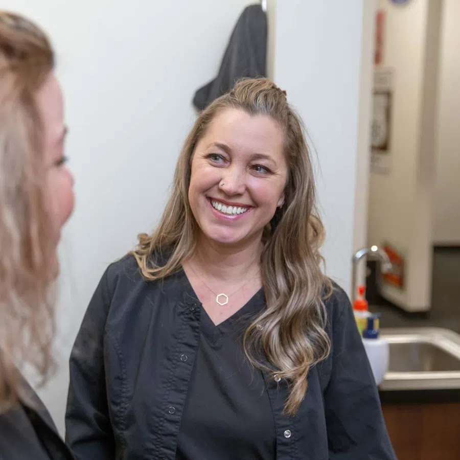 Smiling woman with long hair wearing black scrubs standing in a bright room with a sink and hand sanitizer.