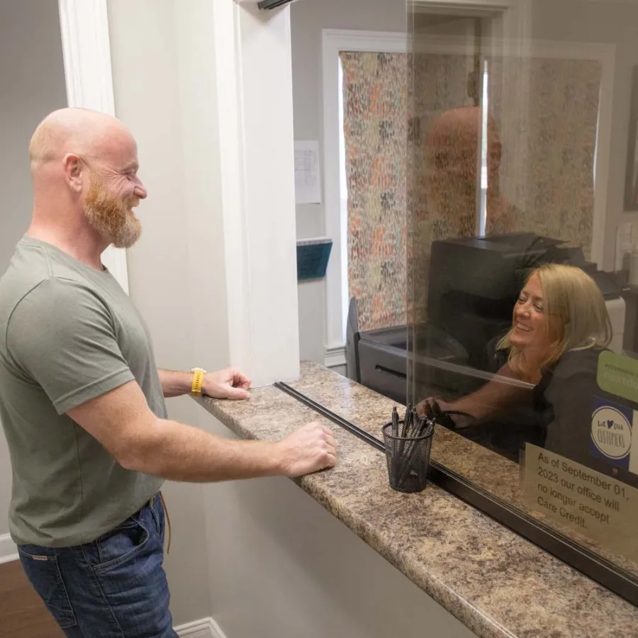Man with beard talks to smiling woman behind protective glass at office reception desk.