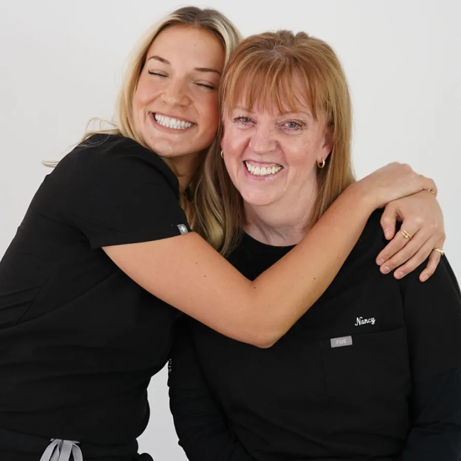 Two women wearing black scrubs smiling and hugging against a plain white background, showing warmth and friendship.