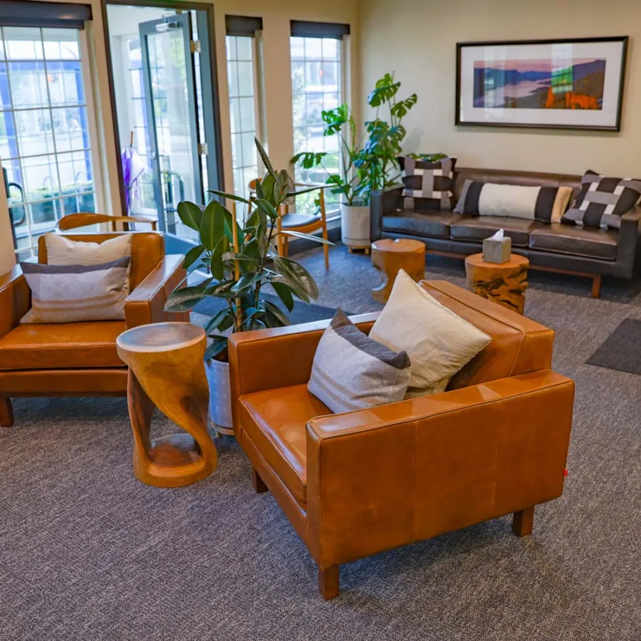 Modern living room with brown leather chairs, plants, gray sofa, wooden tables, and framed artwork on the wall