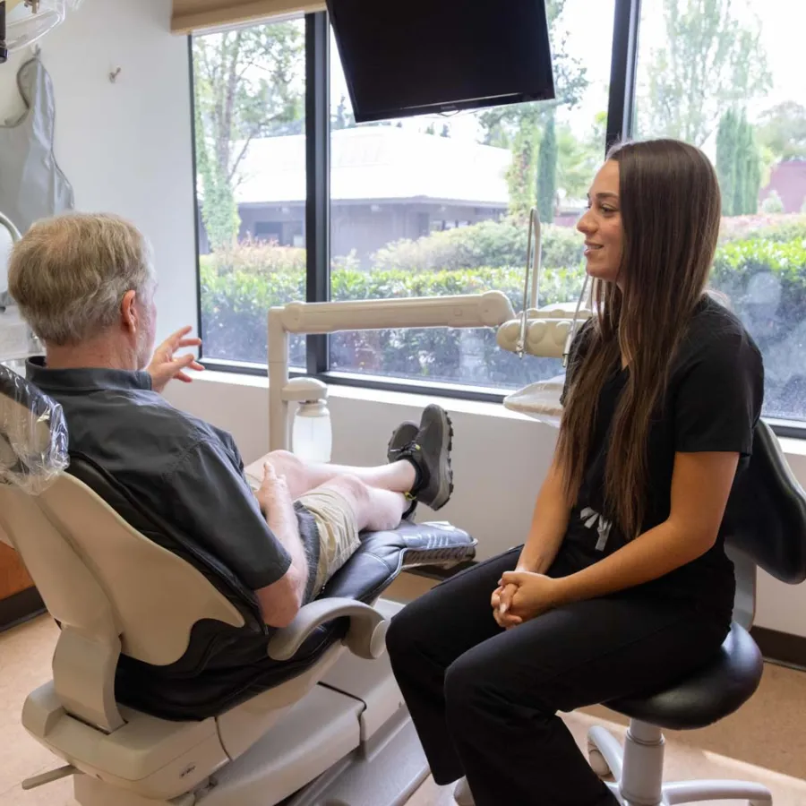 Dentist consulting an elderly male patient in a bright dental office with large windows and modern equipment.