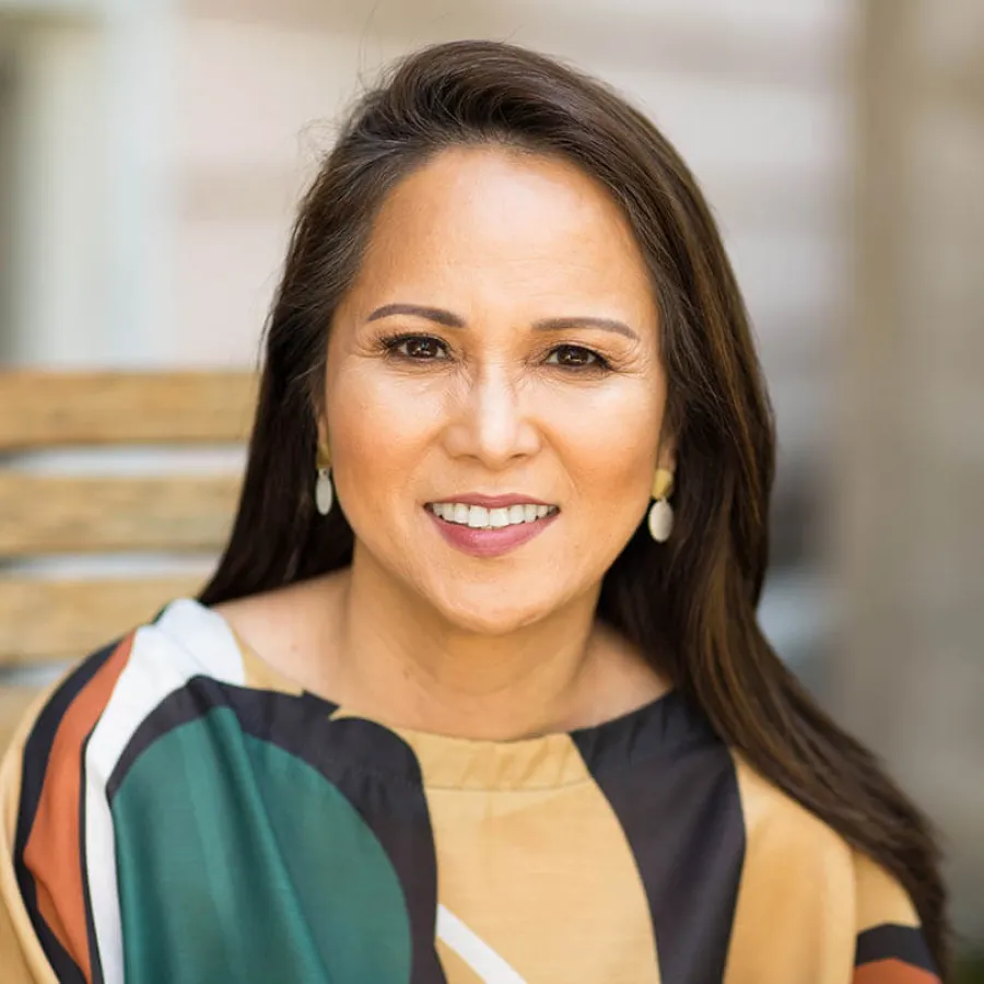 Smiling middle-aged woman with long dark hair wearing colorful patterned top and white earrings outdoors