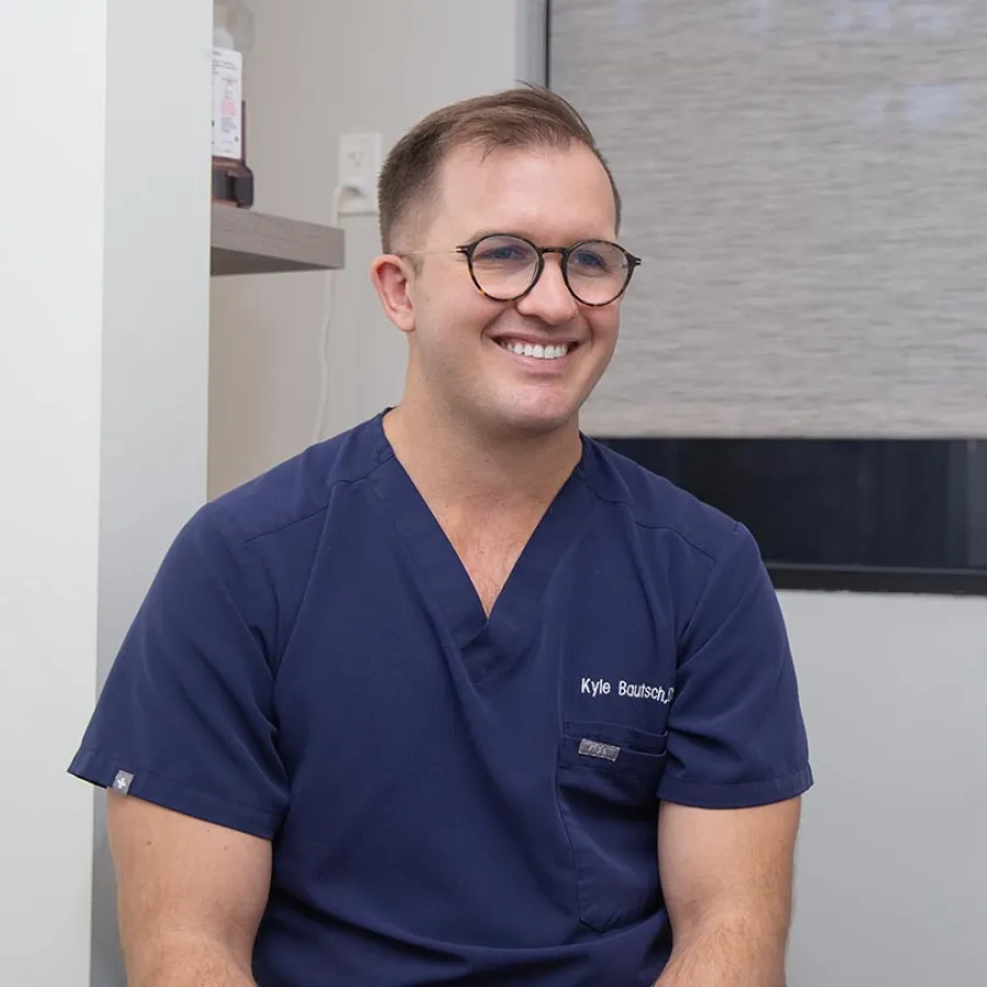 Smiling man in navy scrubs and glasses sitting indoors in a medical or clinical setting.