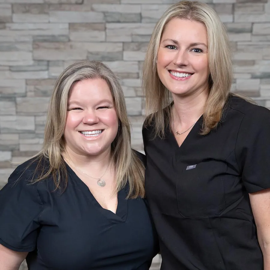 Two smiling women in black medical scrubs standing in front of a stone wall background.