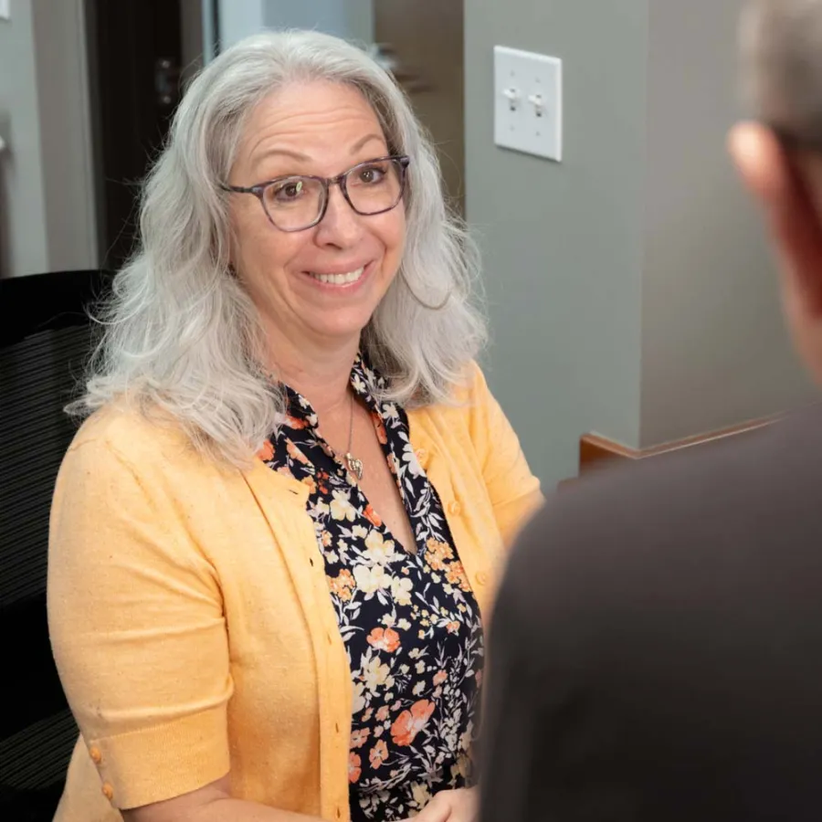 Smiling woman with gray hair and glasses wearing a yellow cardigan talks to a man in an office setting.