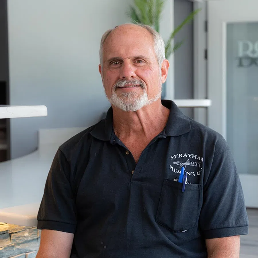 Smiling older man with gray beard wearing a black Strayhart Plumbing polo shirt sitting indoors.