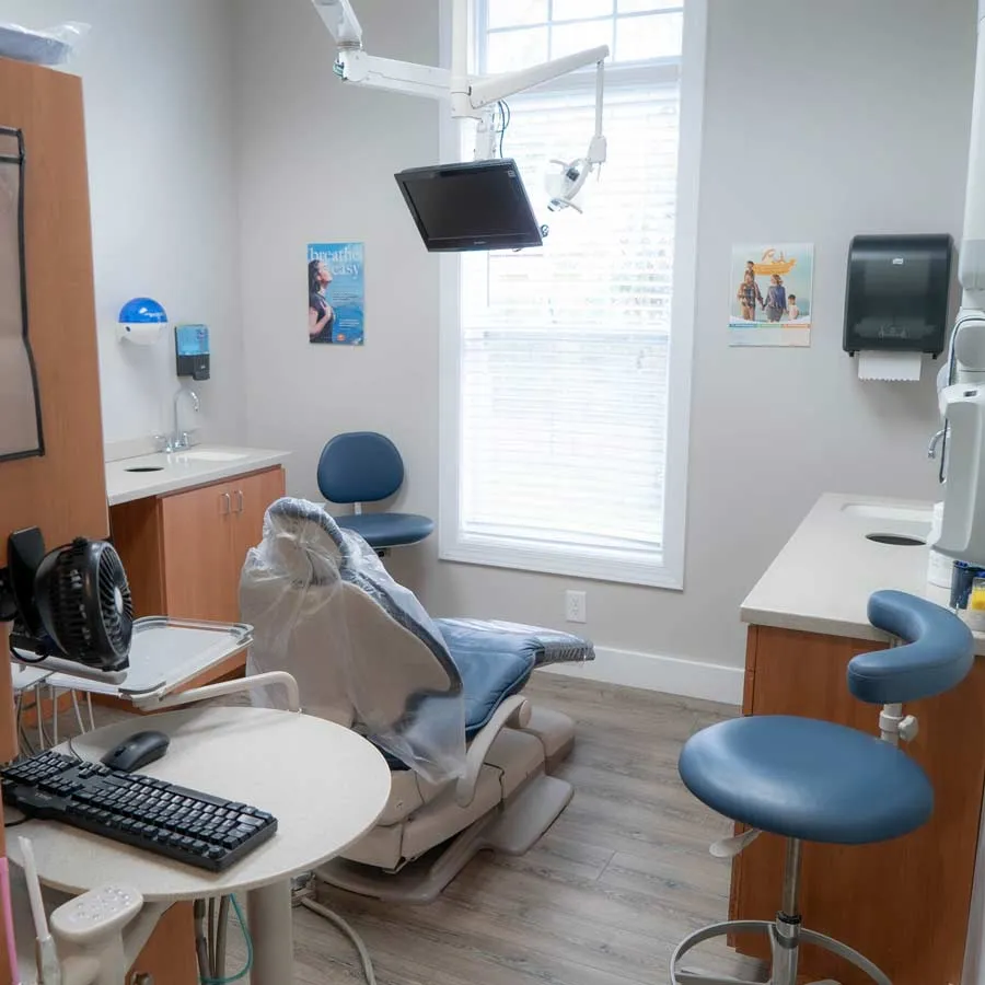 Modern dental exam room with dental chair, computer, stools, and medical equipment under natural light.