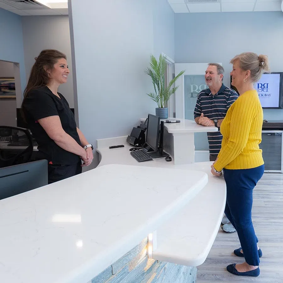 Receptionist talking to an elderly couple at a modern medical office with a white marble counter and indoor plant.