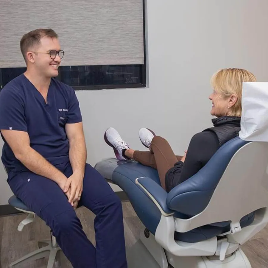 Dentist in navy scrubs smiling and talking with a woman seated comfortably in a dental chair in a modern clinic.