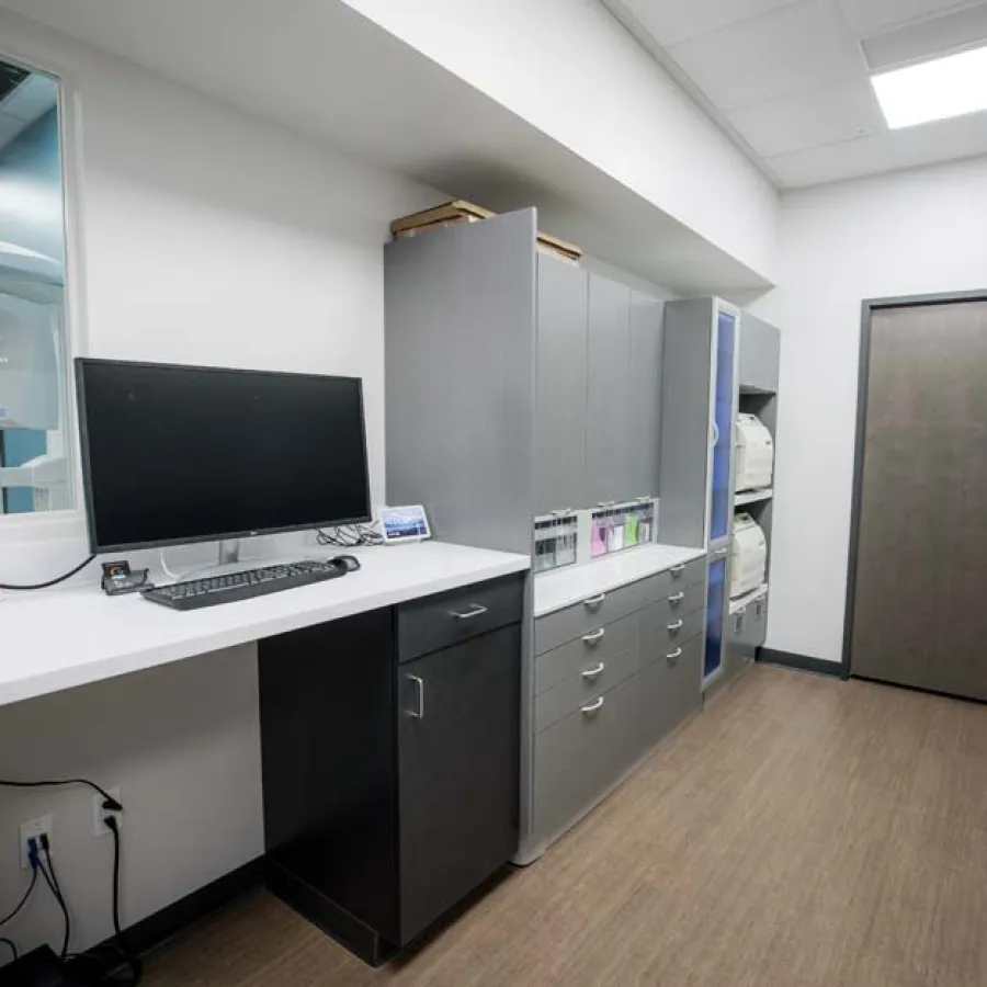 Modern dental office workspace with computer, storage cabinets, and visible X-ray machine through window.