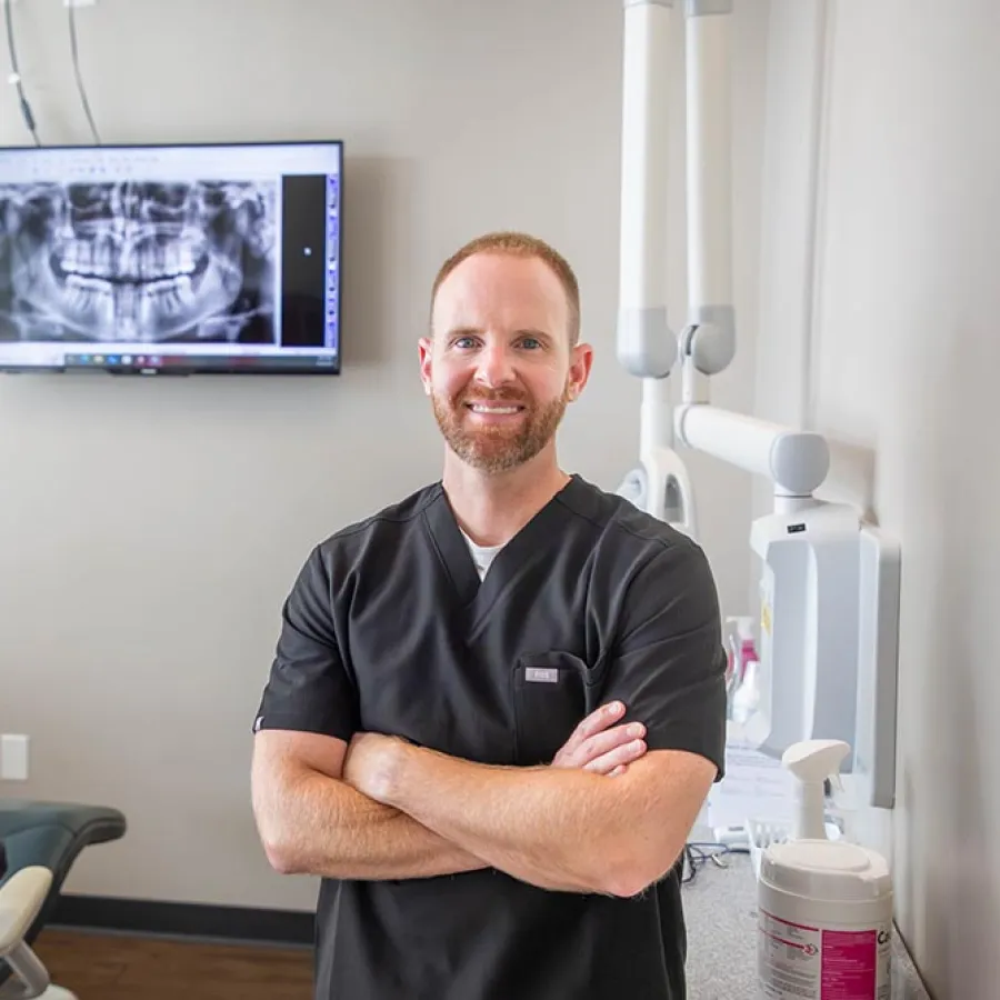 Confident male dentist in black scrubs stands with arms crossed in modern dental office with X-ray on screen.