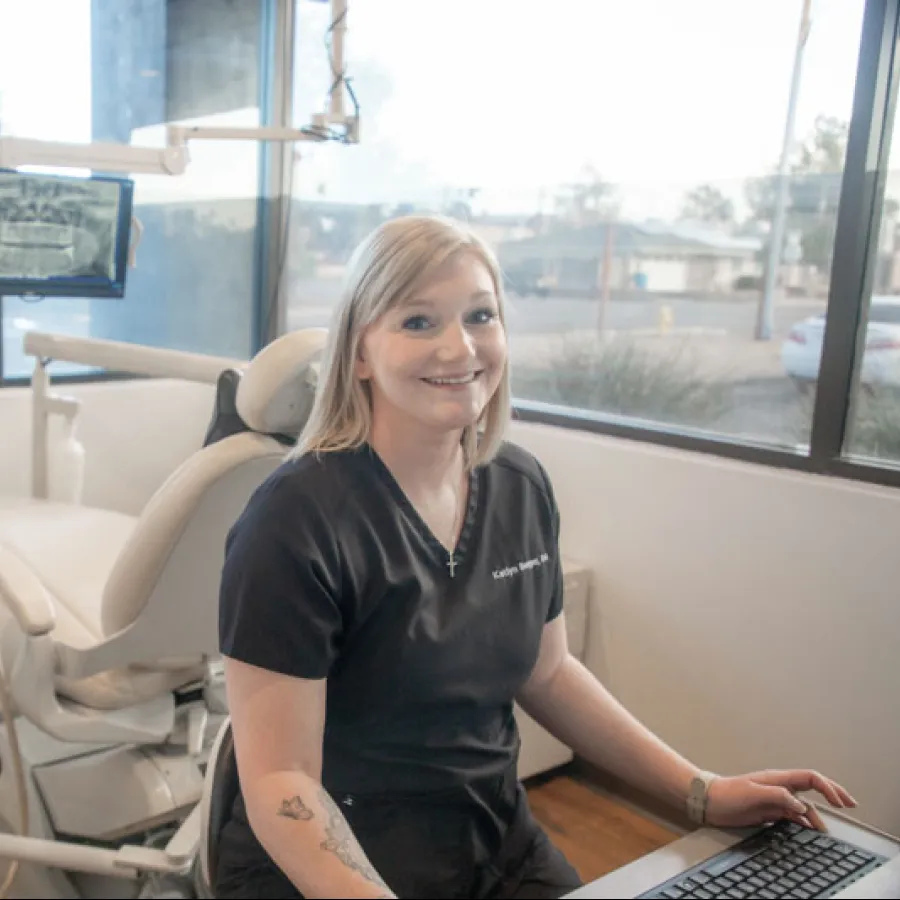 Smiling female dentist in black scrubs sitting at desk in a modern dental office with dental chair and equipment.