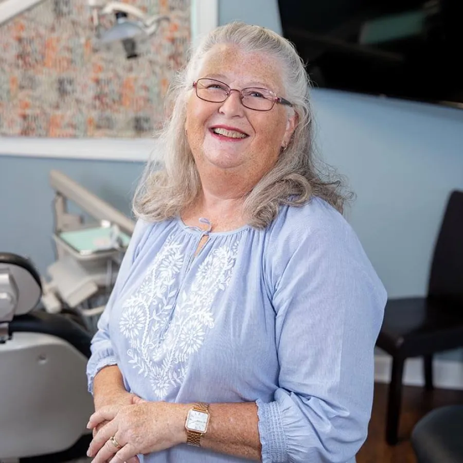 Smiling elderly woman with glasses and gray hair wearing a blue embroidered blouse in a bright room.