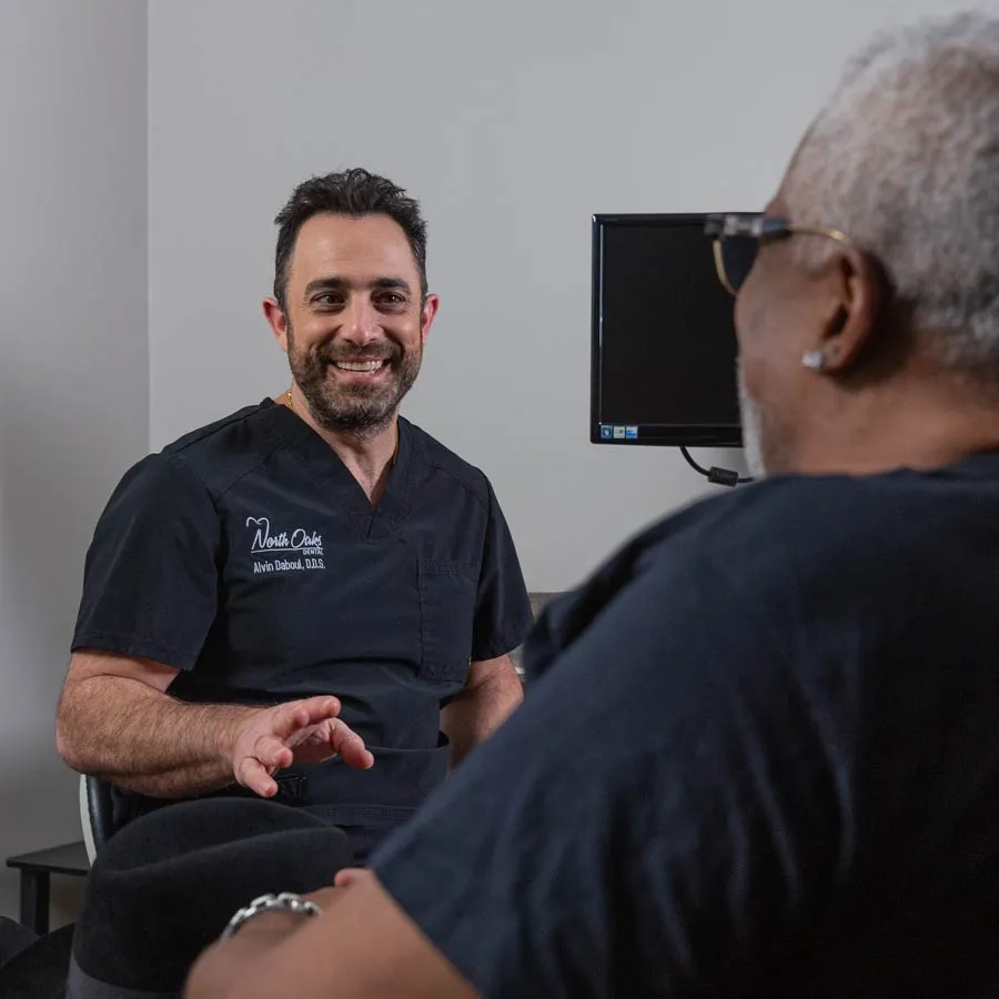 Dentist in black scrubs smiling and talking to an older male patient in a consultation room.
