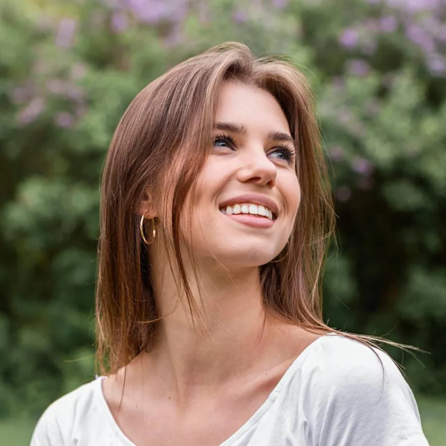 Smiling young woman with brown hair and hoop earrings outdoors with greenery and purple flowers behind her