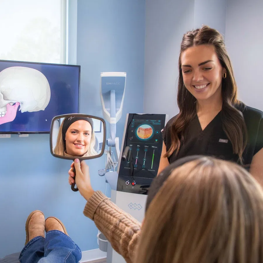Dental patient looks in handheld mirror while dentist explains jaw anatomy with skull images on screen.