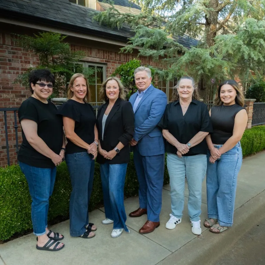 Six diverse adults smiling and standing in a row outside a brick building with greenery on a sunny day.