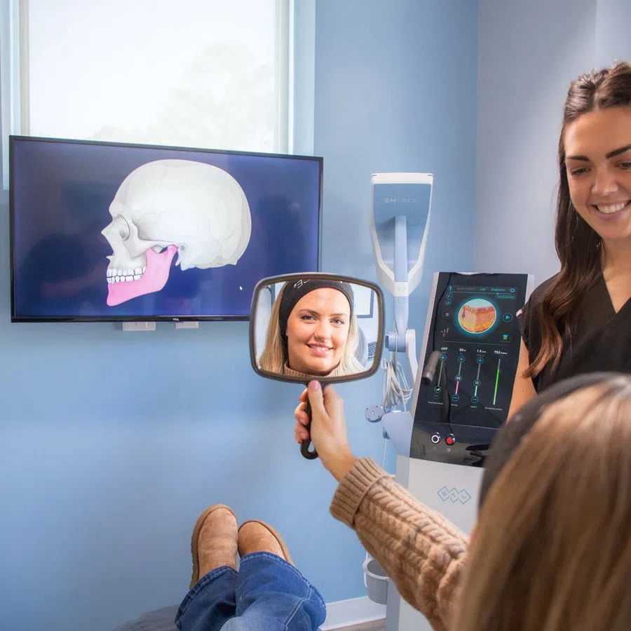 Dental patient looks in handheld mirror while dentist explains jaw anatomy with skull images on screen.