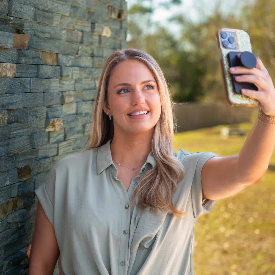 Young woman with blonde hair wearing a light shirt takes a selfie outdoors by a stone wall on a sunny day