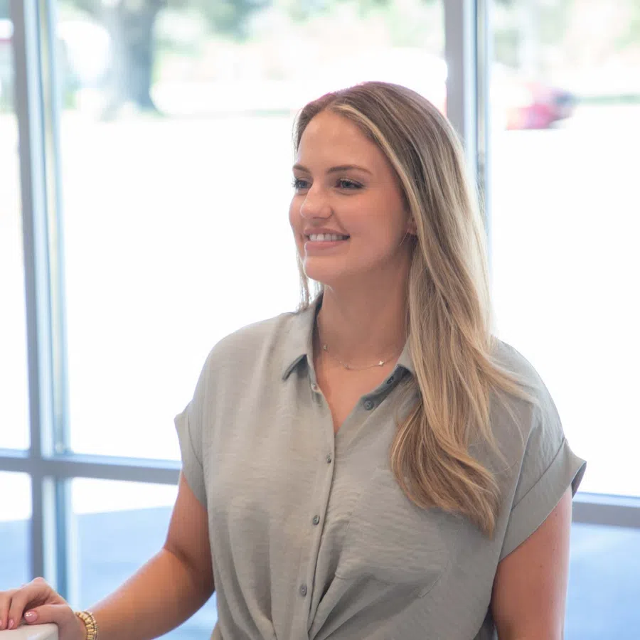 Smiling woman with long blonde hair wearing a grey blouse standing near large windows in bright room.