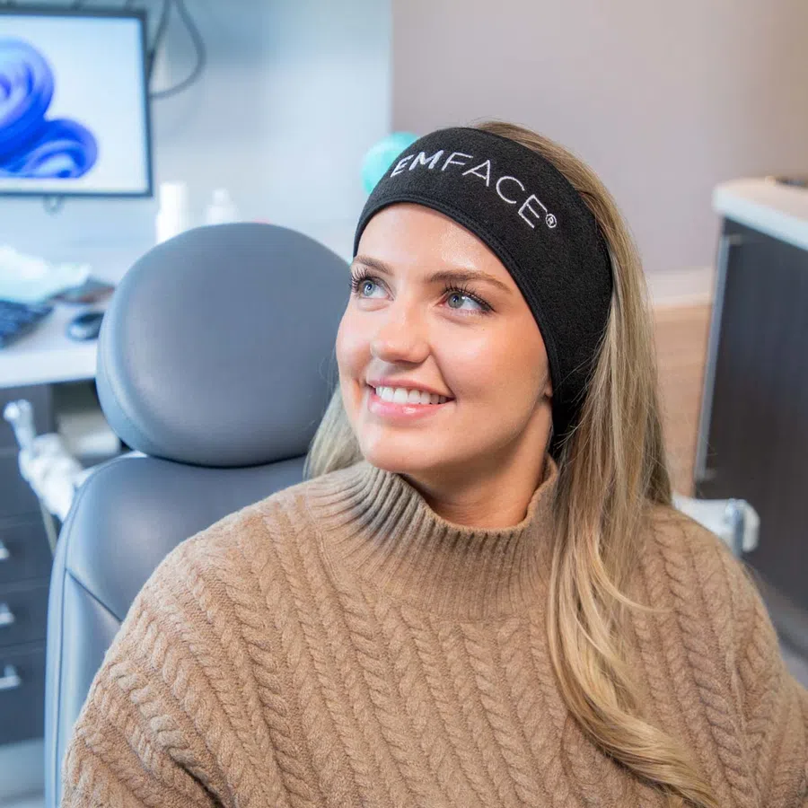 Smiling woman wearing a black EMFACE headband sitting in a modern clinic chair with computer in background