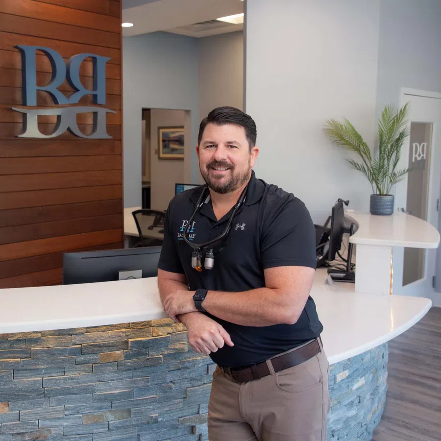 Smiling dentist leaning on modern reception desk with stone facade and plants in a dental office lobby.