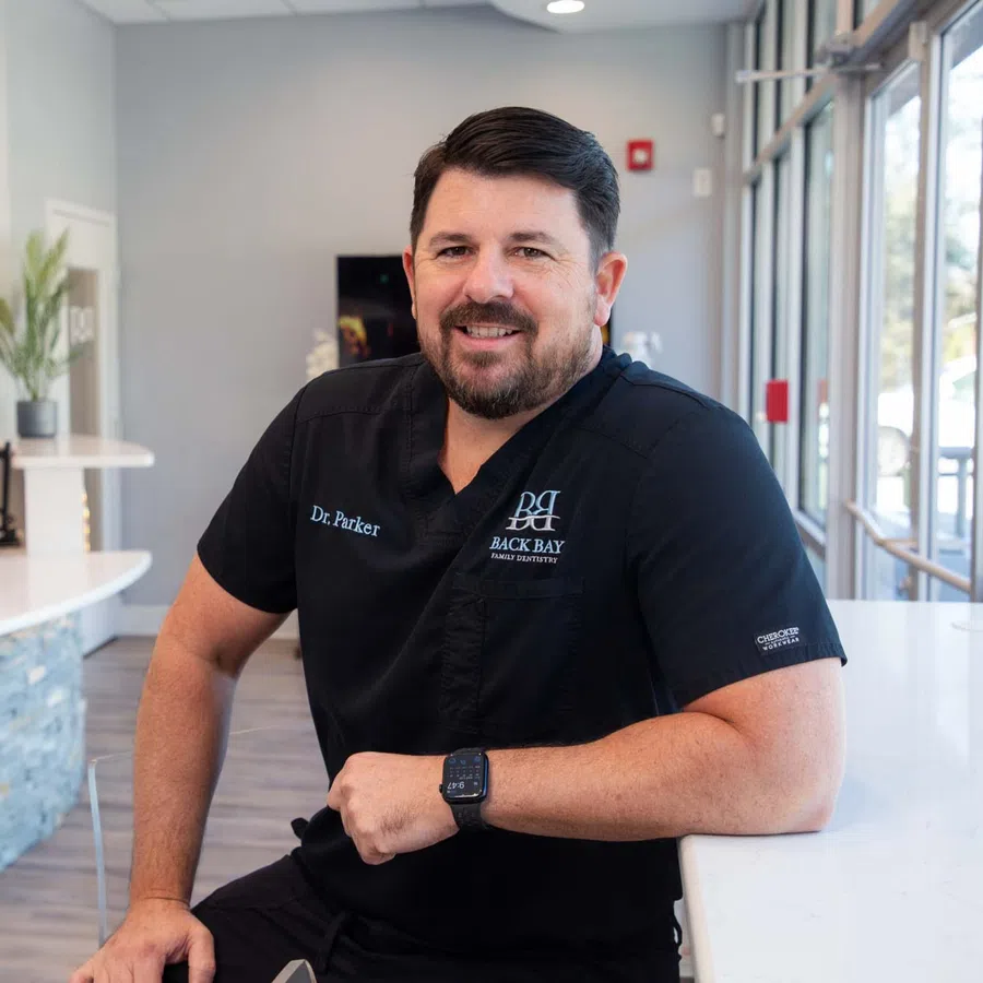 Smiling male doctor in black scrubs leans on white counter in modern medical office reception area