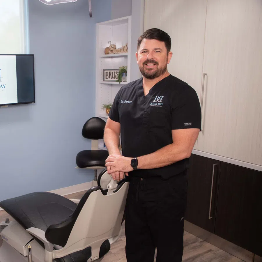 Dentist in black scrubs standing beside dental chair in modern clinic with Back Bay Dentistry logo on screen