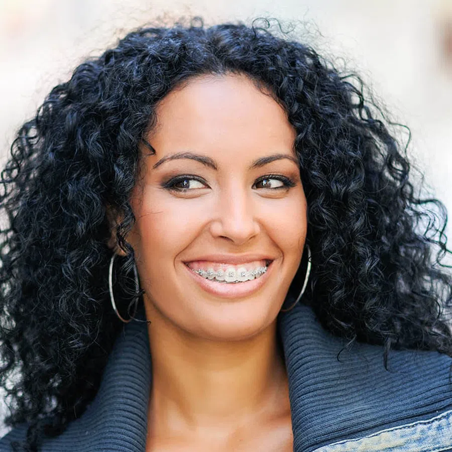 Smiling woman with curly black hair wearing braces and hoop earrings, dressed in a dark jacket outdoors.
