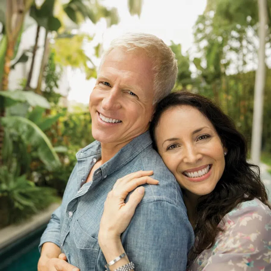 Happy middle-aged couple smiling outdoors with greenery in the background, embracing affectionately by the pool.