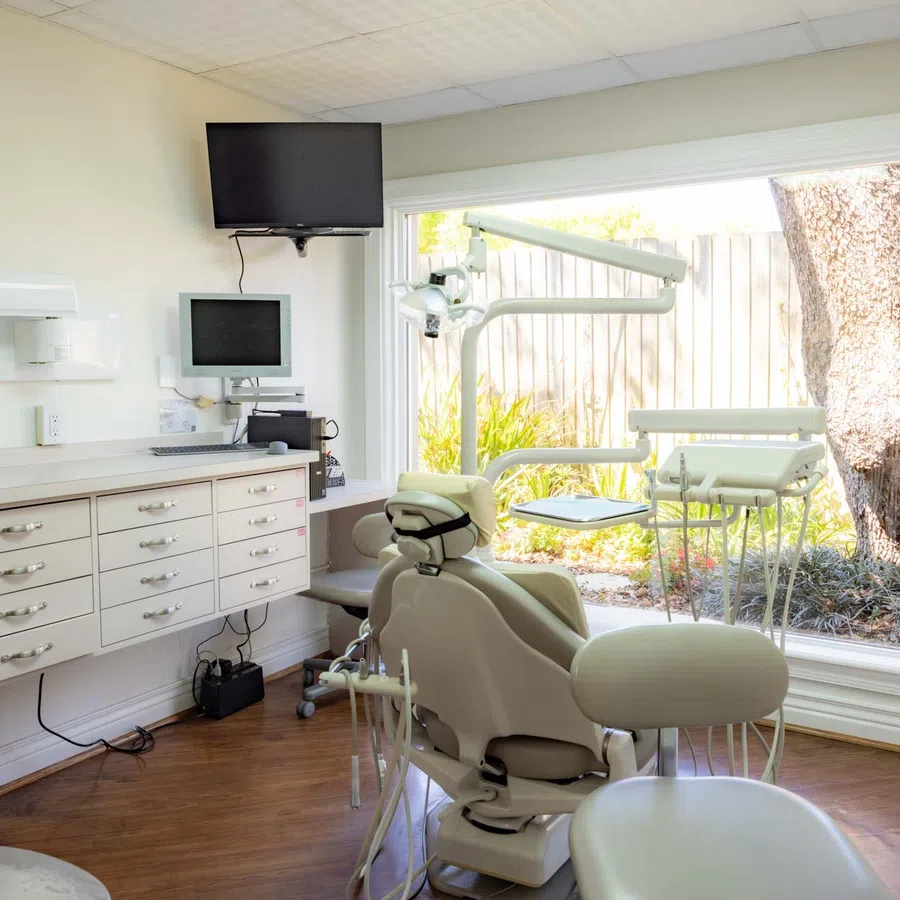 Bright dental office with a patient chair, dental equipment, computer, and large window overlooking garden.