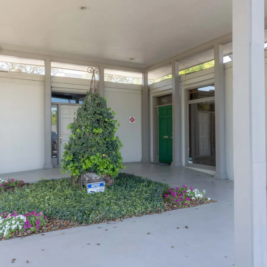 Covered outdoor corridor with green doors, potted plants, and a central landscaped area under white columns.