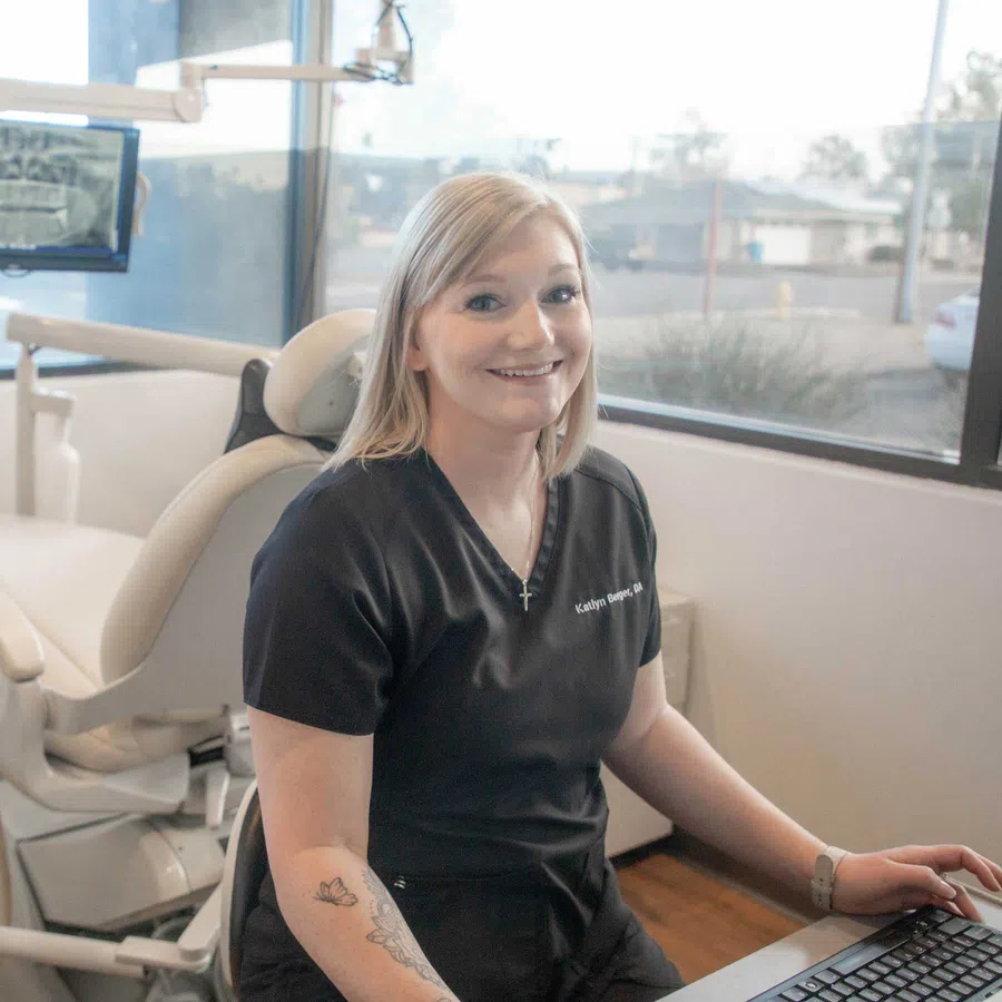 Smiling female dental professional sitting at a desk in a bright clinic with dental chair in background