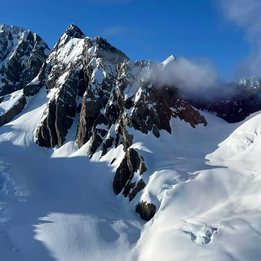Snow-covered jagged mountain peaks under a bright blue sky with light clouds casting shadows